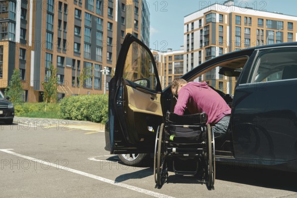 A man moves to a wheelchair from a car parked at a lot for people with disabilities in a contemporary urban parking area. The setting reveals modern buildings in the background under clear skies