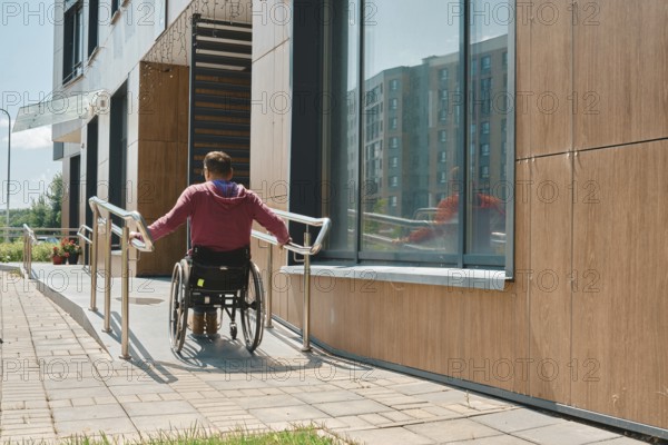 Rear view of a man in a wheelchair is maneuvering up a ramp beside a contemporary building. The clear sky and bright sunlight illuminate the scene, showcasing accessibility features in urban design