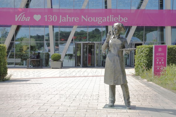 Sculpture confectioner with tray full of pralines for tasting, presentation, eating, enjoyment, pleasure, look, marvel, look at, enjoy, modern, round, building, glass window, pink banner with inscription, anniversary 130 years of nougat love, food, chocolate factory, sale, production and manufacture of sweets, chocolate, nougat, confectionery, confectioner, chocolate, entrance, Viba Nougat World, Schmalkalden, Franconia, Thuringia, Germany