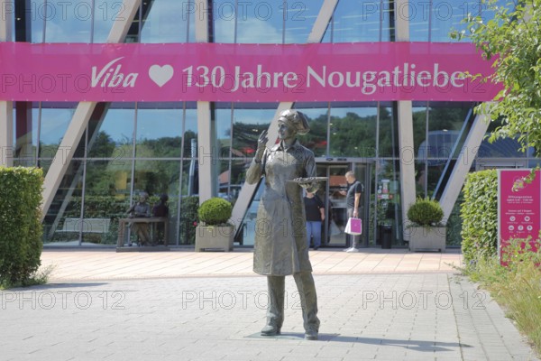 Sculpture confectioner with tray full of pralines to taste, presentation, eat, enjoyment, pleasure, look, marvel, look at, enjoy, modern, round, building, glass window, pink banner with inscription, anniversary 130 years of nougat love, chocolate factory, sale, production and manufacture of sweets, chocolate, nougat, confectionery, confectioner, chocolate, entrance, visitor, Viba Nougat World, Schmalkalden, Franconia, Thuringia, Germany