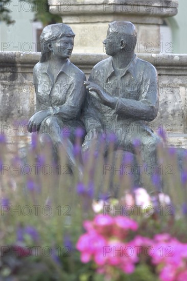 Bronze sculpture at the market fountain, modern art, male figure, female figure, couple, sitting, conversation, entertainment, talking, listening, looking, joy, joyful, friends, relationship, communication, friendship, partnership, floral decoration, depth of field, blur, Schleusingen, Thuringia, Germany