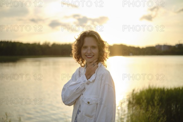 A woman stands by the lake, smiling as the sun sets behind her, casting a warm golden glow on the water. It is a hot day, perfect for enjoying the scenery, nature, and tranquility