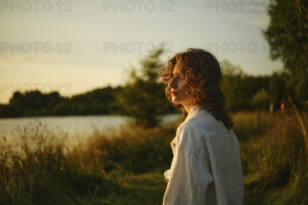 On a bright, hot day at the lake, a young person stands beside the water, absorbed in the stunning sunset. Tall grass sways gently as the sun casts a warm glow, creating a peaceful atmosphere