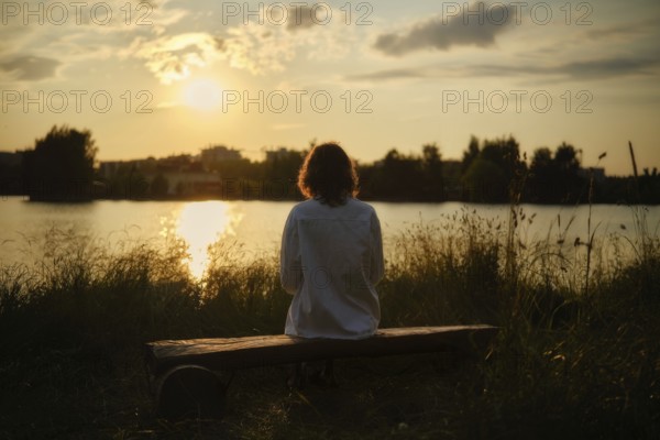 At the edge of a serene lake, a woman sits on a wooden bench surrounded by tall grass, soaking in the warm sun and vibrant colors of the sunset