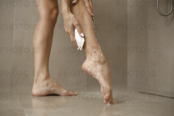 A woman is focused on washing her leg in a bathroom. The soft natural light enhances the clean setting, with water droplets glistening on her skin and the tiled floor