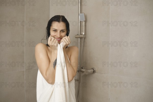 A woman with wet hair smiles while holding a towel in a modern bathroom. The tiles are light, and a showerhead is visible in the background, creating a relaxing atmosphere