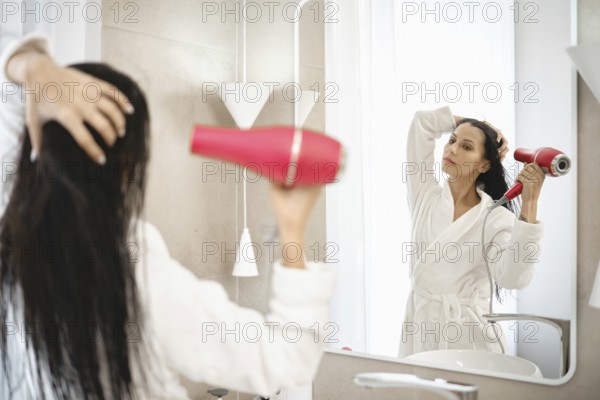 A woman is using a hair dryer to style her wet hair while standing in front of a large mirror. The bright bathroom features minimalistic design elements and soft natural light filtering through