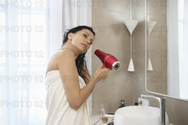 A woman stands in a well-lit bathroom, wrapped in a towel and using a vibrant red hair dryer to dry her wet hair