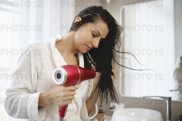 A woman wearing a white bathrobe is focused on drying her long, wet hair with a red hairdryer. The bathroom is well-lit, featuring a large mirror and modern fixtures