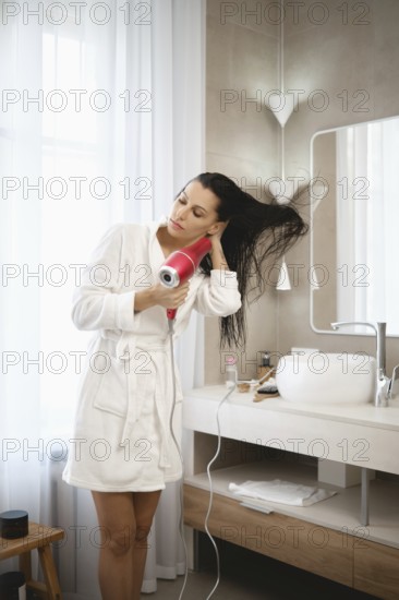 A woman stands in a modern bathroom, using a hair dryer to style her long, wet hair. She is dressed in a cozy white robe, surrounded by minimalist decor, preparing for her day