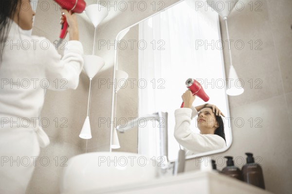 A woman wearing a bathrobe uses a hairdryer while looking in the mirror. The chic bathroom features elegant lighting, minimalistic design, and various hair care products on display