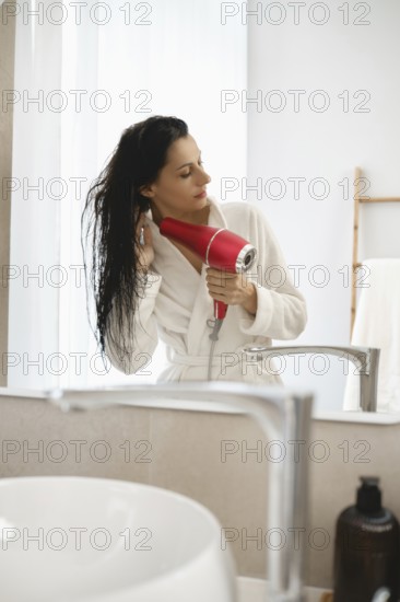 A woman in a white robe stands in a well-lit bathroom, carefully drying her wet hair with a bright red hairdryer. The mirrors reflect the serene setting, adding to the relaxing atmosphere
