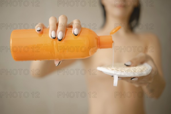 A person with wet hair uses an orange bottle to dispense liquid soap onto a textured sponge. The setting is minimalistic and modern, focusing on personal care and hygiene routines