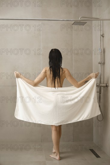 A woman stands in a shower holding a large white towel. Water droplets linger on her wet hair. The backdrop features neutral tiles, enhancing the calm and clean atmosphere of the space