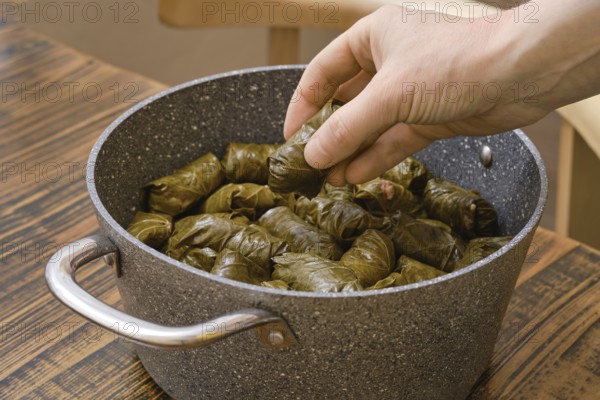 A hand carefully adds stuffed grape leaves to a pot filled with dolma in a warm home kitchen. The table has a rustic wooden finish, creating a welcoming atmosphere for cooking and sharing