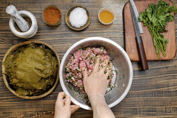 At home, a person mixes seasoned minced meat with rice and herbs in a large bowl. Surrounding the workspace are bowls of spices, vine leaves, and chopped greens ready for making dolma