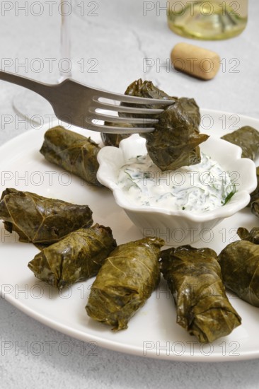 Dolma is being prepared in a cozy kitchen. Stuffed grape leaves are arranged on a plate, accompanied by a small bowl of yogurt sauce for dipping. The culinary process is evident and inviting