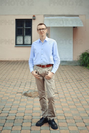 A man stands confidently outside a modern building, wearing a light blue shirt and beige pants. His dark shoes complement the outfit