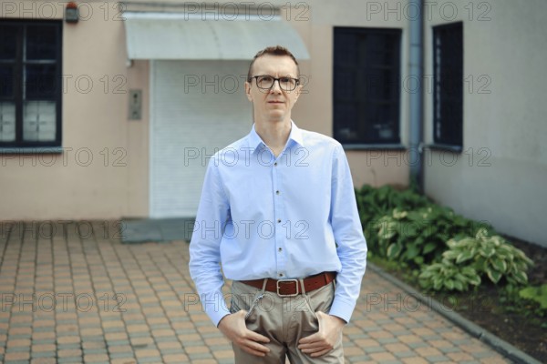 A man with glasses in a light blue shirt and beige trousers standing in front of a building. He has a relaxed posture with hands in pockets, surrounded by greenery and bricks