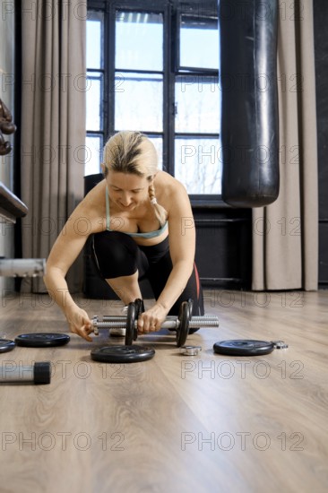A focused woman is adjusting weights while kneeling on a gym floor. The space has large windows allowing natural light to illuminate the workout area, creating an inspiring atmosphere