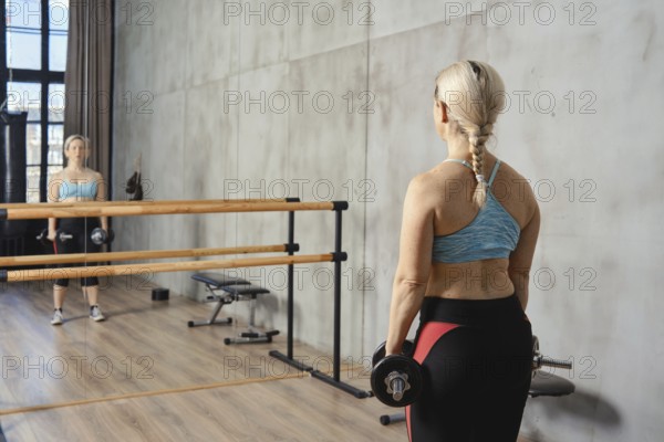 A senior woman stands in front of a mirror in a contemporary gym, holding dumbbells. She is focused on her reflection while engaging in a strength training workout to enhance her fitness