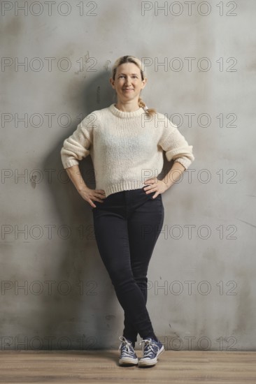 A woman stands casually with her hands on her hips, wearing a soft sweater and dark jeans. She smiles softly, exuding confidence against a muted wall in a well-lit indoor setting