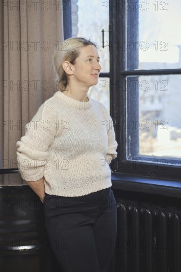 A woman with light hair is standing by a window, gazing thoughtfully outside. She is dressed in a cozy cream sweater and dark pants, surrounded by warm sunlight filtering through the glass