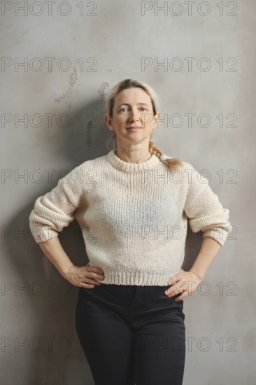 A woman with long, light hair stands upright with her hands on her hips, wearing a soft beige sweater and dark pants. She exudes poise in front of a light gray wall, showcasing calm confidence