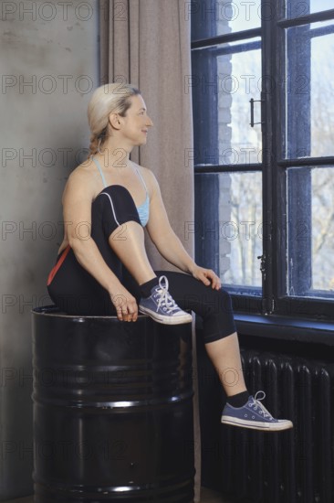 A woman sits on a barrel, pausing between exercises. She gazes out the large windows, showcasing her focus and serenity in the tranquil environment