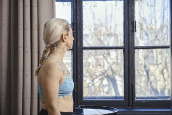 A woman with braided hair is standing near a large window, looking outside at the bright day. She wears a blue athletic top and appears thoughtful, surrounded by soft curtains and natural light