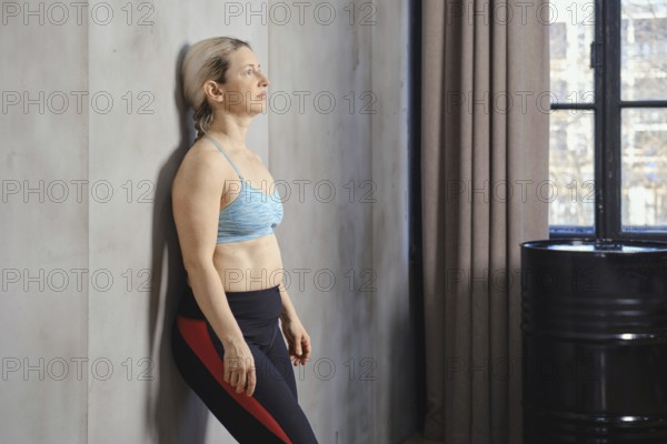 A woman leans against the wall in a modern studio, focusing on her breath as sunlight filters through the window. She wears athletic attire, embodying calm and relaxation during her practice