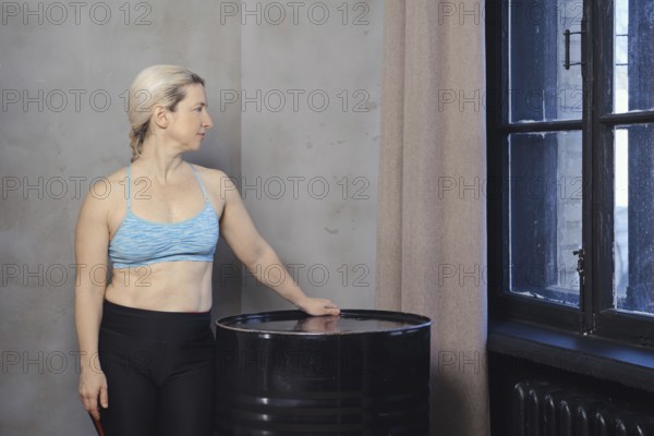 A woman wearing a blue sports bra and black leggings stands next to a black barrel, gazing thoughtfully out the window. The minimalist room features a neutral-toned wall and soft natural light