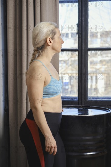 A woman stands near a window, wearing a sports bra and leggings, looking thoughtfully outside. The room is well-lit, highlighting her focused expression as she engages in a morning routine