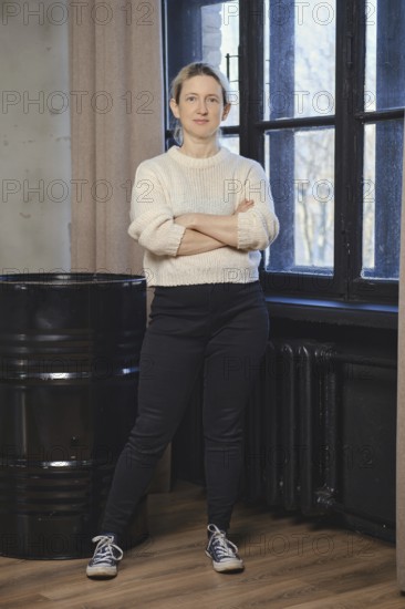 A woman stands with arms crossed, dressed in a white sweater and dark pants, exuding confidence in a well-lit indoor setting. Natural light streams through the window, illuminating the space