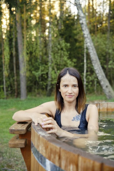 Outdoor portrait of pretty woman in a wooden hot tub surrounded by lush greenery. The warm glow of the evening sun enhances the serene atmosphere, creating a perfect setting for relaxation