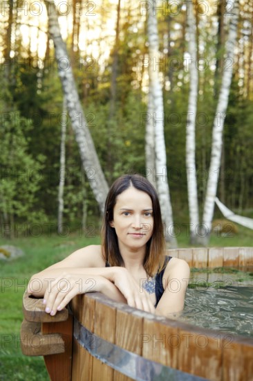 A woman enjoys a tranquil moment in a wooden hot tub, framed by tall trees and lush greenery during sunset. The warm light adds a calming ambiance to her peaceful retreat