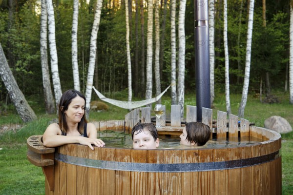 A woman and two children share a joyful moment in a wooden hot tub surrounded by trees. The afternoon sun creates a serene atmosphere, while a hammock hangs in the background for lounging