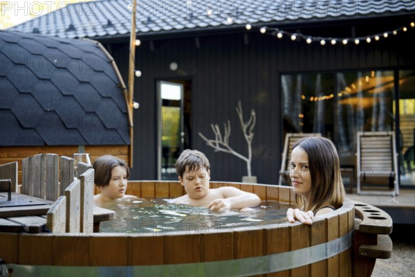 Mother and two children relax in a wooden hot tub in a backyard. The warm water scented with fir branches has a calming effect and relaxes the body