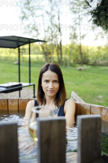 A woman enjoys a serene moment in a wooden hot tub, immersed in warm water, while surrounded by lush greenery. Bright sunlight filters through the trees, creating a peaceful atmosphere