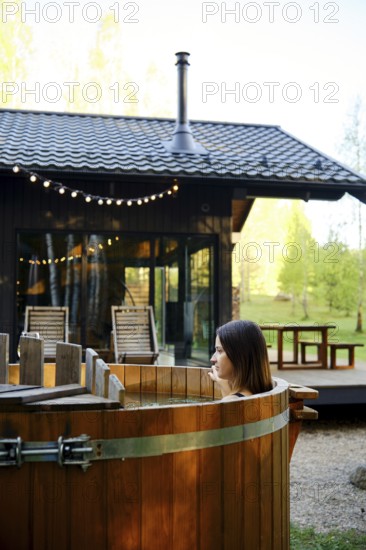 A woman enjoys relaxing in a wooden hot tub in the backyard of a tiny house. The sun illuminates the surrounding greenery, and warm lights add a cozy ambiance to the tranquil setting