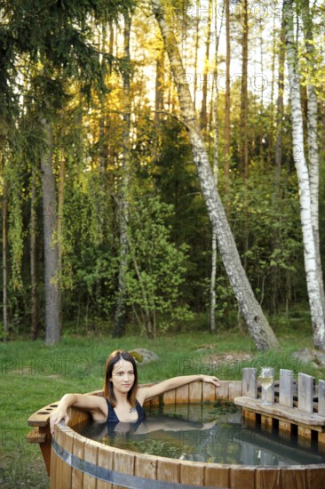 A woman relaxes in a wooden hot tub nestled in a forested area. The warm sunlight filters through the trees, creating a tranquil atmosphere perfect for relaxation