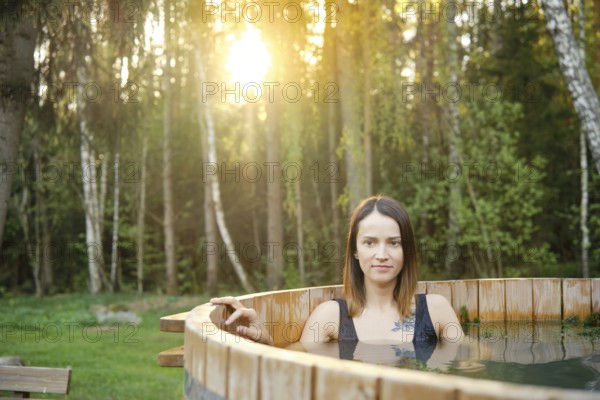 A woman enjoys a soothing soak in a wooden hot tub amidst tall trees during a serene sunset. The warm light filters through the leaves, creating a peaceful and calming atmosphere