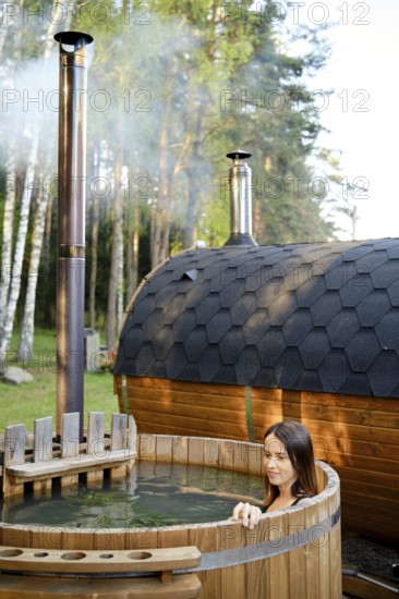 A woman enjoys a peaceful moment in an outdoor wooden hot tub surrounded by tall trees. Steam rises from the sauna, creating a calm atmosphere perfect for relaxation in nature