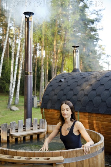 A woman enjoys a peaceful moment in a wooden hot tub, soaking in warm water. The nearby sauna sends steam into the air as trees provide a beautiful backdrop under a clear blue sky