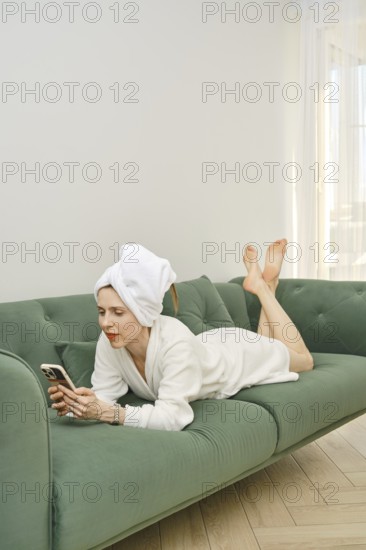 A woman in a white bathrobe and towel is lounging on a green couch, scrolling on her smartphone. Natural light filters through the nearby window, creating a serene atmosphere perfect for relaxation