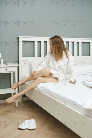 A woman wearing a white robe sits on the edge of a neatly made bed in a minimalist bedroom. She rubs the cream into her leg with a thoughtful expression on her face