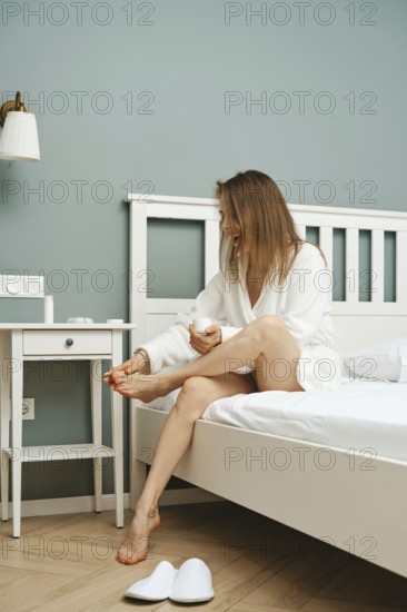 A woman in a white robe smiles while applying lotion to her feet in a charming bedroom. Natural light falls on the bed and bedside table, creating a relaxing morning atmosphere