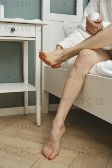 A person sits on the edge of a bed, applying cream to her foot while enjoying a moment of self-care in a cozy bedroom. Soft lighting and calm colors create a soothing atmosphere
