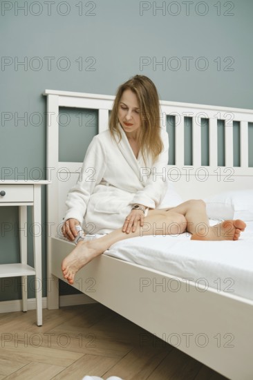 A woman in a white bathrobe peeling dead skin with electric exfoliating machine sitting in bedroom