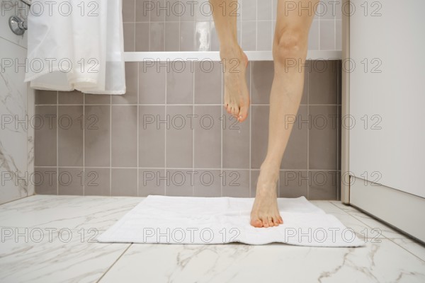 A person is stepping onto a white towel placed on the floor of a sleek bathroom after taking a shower. Water is dripping from her foot onto the towel, highlighting cleanliness and daily routines